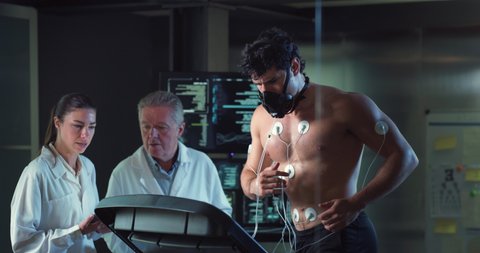 Cinematic shot of young athlete with medical electrodes doing endurance test of body resistance by training on treadmill while physicians monitoring heart rate, pulse and ECG data in modern laboratory