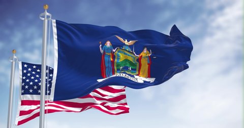The New York state flag waving along with the national flag of the United States of America. In the background there is a clear sky. New York is a state in the Northeastern United States