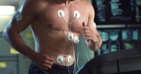 Cinematic close up shot of young athlete with medical electrodes doing endurance test of body resistance by training on treadmill with monitoring heart rate, pulse and ECG data in modern laboratory.