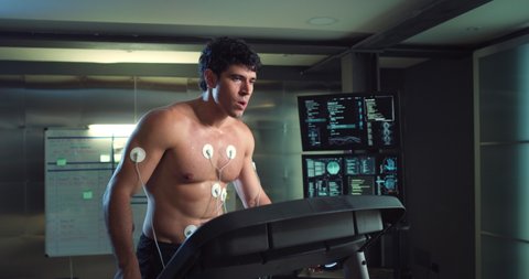 Cinematic shot of young athlete with medical electrodes doing endurance test of body resistance on treadmill and using futuristic screen for graphic implementation in modern laboratory.