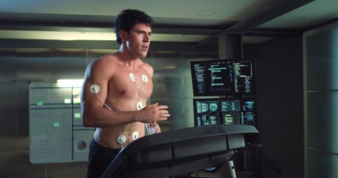 Cinematic shot of young athlete with medical electrodes doing endurance test of body resistance on treadmill and using futuristic screen for graphic implementation in modern laboratory.