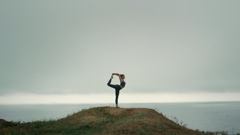 Fit woman standing one leg stretching on hill cloudy day. Sporty girl doing gymnastic exercise holding foot in hand outdoors. Attractive flexible lady practicing yoga natarajasana on beach hilltop. 