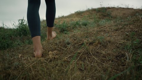 Athletic woman legs walking up hill wearing dark leggings close up. Unknown sporty girl climb hilltop on green grass outdoors. Bare feet stop in front calm grey ocean cloudy day. Nature concept.