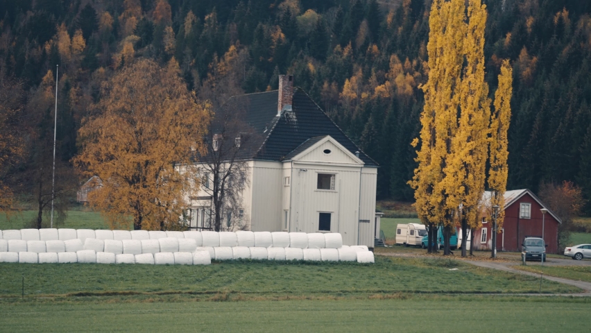 A farm in rural Norway. Hay bales are stacked in the field near the house. Cars are parked nearby. A person walks to the house. Slow-motion.