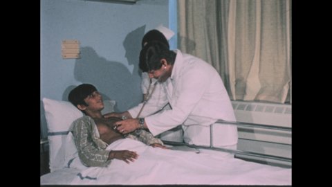 1970s: Doctor examines young patient laying in hospital bed. Doctor gives stethoscope to patient. Boy listens through stethoscope, smiles.