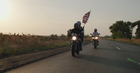 A group of young men rides motorcycles at sunset, followed by the U.S. flag