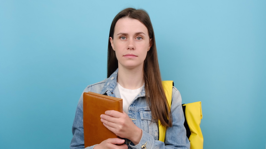 Portrait of girl teen student wears denim jacket and yellow backpack hold books say oops ouch oh omg, posing isolated over blue color background. Education in high school university college concept Royalty-Free Stock Footage #1090050507