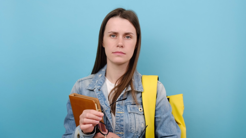 Tired girl teen student hold books glasses rub nose, smiling looking at camera, wear denim jacket and yellow backpack, isolated on blue background. Education in high school university college concept