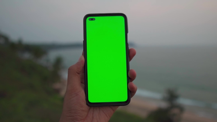 POV shot of hand holding mobile with green screen in front of the sea during the sunset. Chroma key mock-up on smartphone in hand at the sea shore in Goa, India. Green screen mockup template of Mobile | Shutterstock HD Video #1090053933