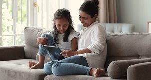 Indian mother little daughter junior schoolgirl cuddle on comfy sofa play internet video game using electronic touchpad. Two diverse age females mom child girl scroll social media pages on tablet pc