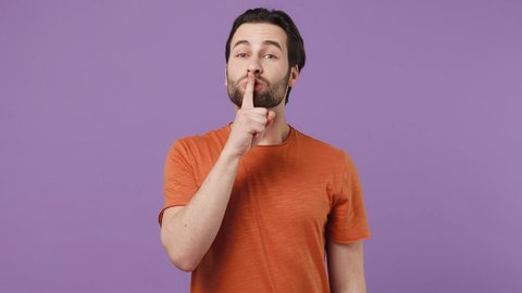 Secret serious fashionable young brunet bearded man 20s wears red t-shirt look aside say hush be quiet with finger on lips shhh gesture isolated on plain pastel light purple background studio portrait