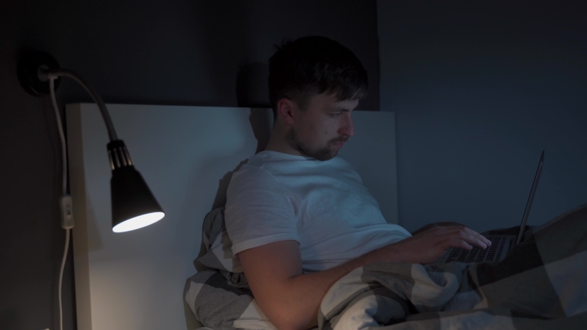 Young caucasian guy waking up at night sitting on bed looking at computer screen with great concentration. Man cannot fall asleep again and starts working on laptop. Insomnia and sleep problems. | Shutterstock HD Video #1090059757