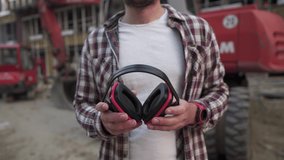 Builder in front of construction machinery puts hearing protection on ears. Worker at construction site in safety goggles and ear protection is proudly posing against heavy equipment excavator. - Powered by Shutterstock - Get 15% off with code: PIKWIZARD15