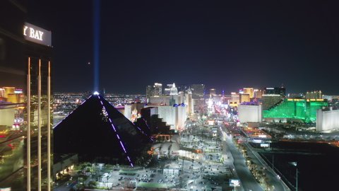 Las Vegas at night, Nevada, Apr. 2022. Cinematic aerial of epic shiny golden building of Mandalay Bay luxury resort and breathtaking Luxor hotel Pyramid building with bright beam in the night sky 4K