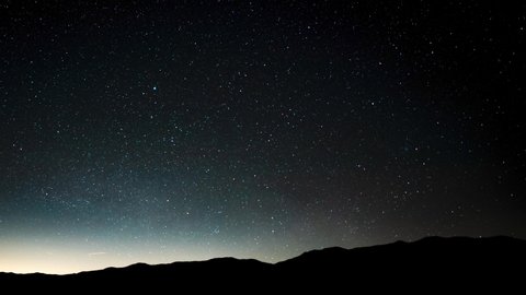 A night sky full of stars and the Milky Way crossing the sky above the horizon of the desert in silhouette - time lapse