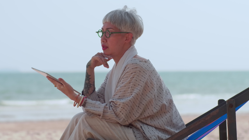 Asian elderly woman using a tablet computer while sitting on the beach.