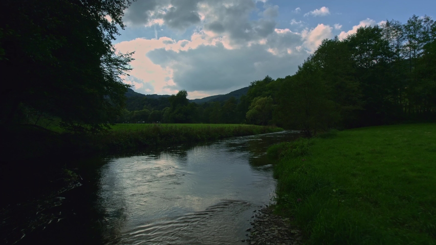 Low angle view over a peaceful calm river Sauer and low underneath the trees at dusk, meadows with fresh green grass, forest surrounding the meadow and a blue sky in the background, in Luxembourg