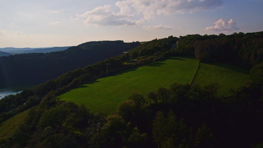 Aerial view: At the top of the mountain a road with cars passing. A meadow with freshly mowed grass, electricity pylons and cables. With mountains and rolling hills in the background in Luxembourg