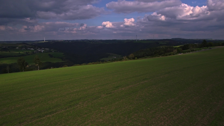 Beautiful landscape aerial view over the mountains of Luxembourg with meadows and a provincial road with two passing cars and in de background the woods with three wind turbines and a blue purple sky
