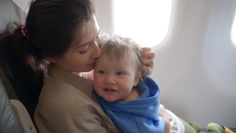 Mother looks out an airplane window holding a baby. Toddler sleeping inside airplane, parent masked holding infant toddler asleep while flying in economy class. Baby sleeping inside airplane, parent