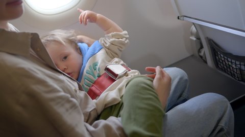 Baby sleeping inside airplane, parent masked holding infant toddler asleep while flying in economy class. young boy sitting seat looking out airplane window while flying. Mother looks out an airplane