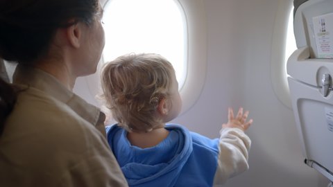 young boy sitting seat looking out airplane window while flying. Baby sleeping inside airplane, parent masked holding infant toddler asleep while flying in economy class. Mother looks out an airplane