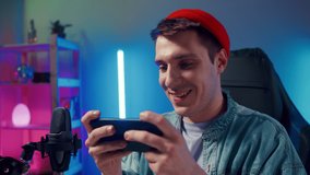Happy guy man gamer wear red hat competition play video game online with smartphone colorful neon light in living room at night modern house. Esport streaming game online, Home quarantine activity.