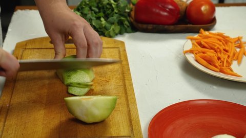 Close-up of a woman's hands taking a green radish off a plate and placing it on a kitchen board. The cook is cutting the green radish with a knife. There are vegetables on the table