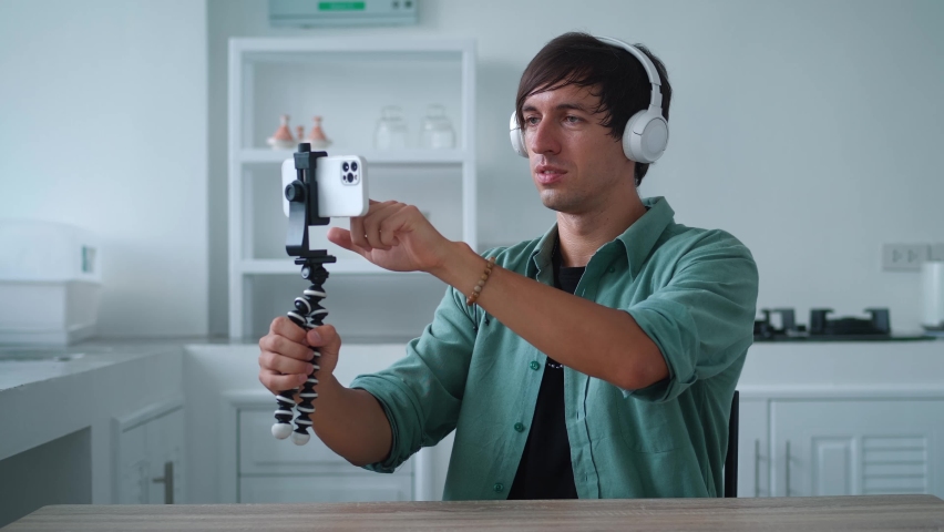 Young man in wireless headphones is talking on a video call using his mobile phone. Popular blogger is recording a video blog for the internet using his smartphone, at home in the kitchen