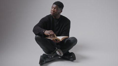 Focused interested black guy wearing black clothes sitting cross-legged on the floor reading a book, turning pages, looking up. High quality 4k footage