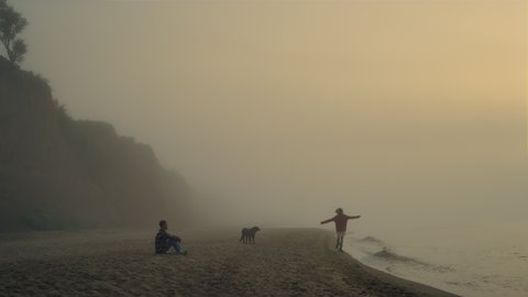 Young couple spending leisure time on beach. Wide shot attractive girl and guy enjoying sunrise landscape at foggy sea. Happy woman dancing at seaside. Relaxed man sitting on shore. Lifestyle concept