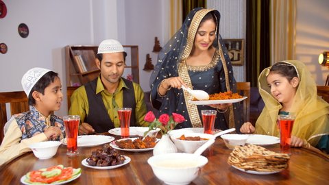 A modern nuclear Muslim family having food together while sitting at the dining table - Eid, festive season. A beautiful housewife in traditional wear serving food to her family members - delicious.