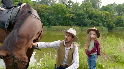 Cowboy and his daughter with their horse on the river bank. Happy cowboy family. Slow motion.
