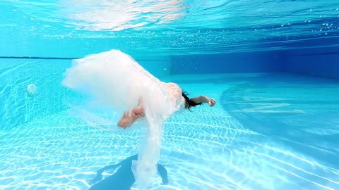 The bride in a white wedding dress swims in the pool underwater. The water is clear and blue. In love