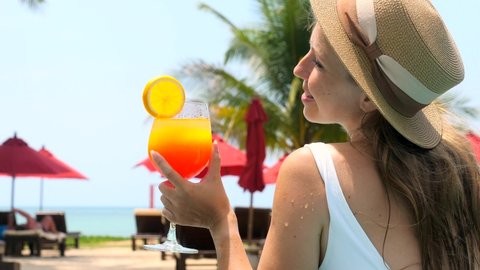 Beautiful girl in swimwear drinking colorful yummy cocktail at swimming pool. Woman in white swimsuit drinking non-alcoholic orange cocktail, relax in pool water, enjoy vacation at luxury hotel