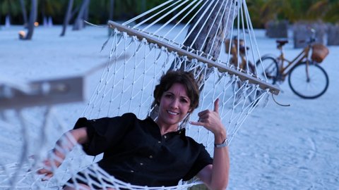 A happy woman makes the shaka sign while relaxing in a hammock, Maldives 