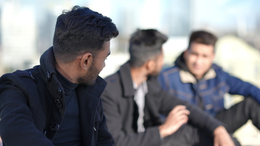 Portrait of handsome young man looking at camera smiling as blurred friends talking at background. Confident Middle Eastern guy posing outdoors partying with mates on rooftop in urban city