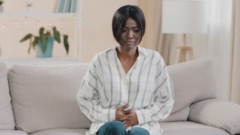 Young upset unhealthy sad african american woman sitting in room on couch holding hands on stomach suffering from abdominal pain feeling spasm from diarrhea menstrual painful feelings symptom nausea