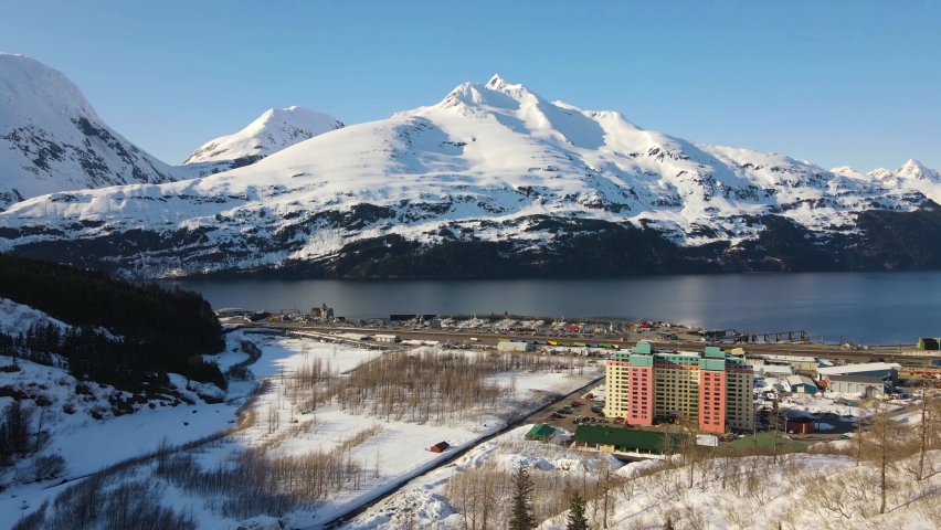 4K cinematic drone view over the town of Whittier Alaska showing a tall rise apartment building with large whitecapped mountains in the background during winter
