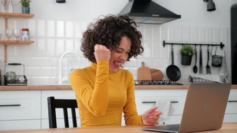 Excited curly woman student winner celebrating admission to university, uses laptop at home kitchen. Happy female sitting on chair at table reads e-mail about college admissions, rejoices victory