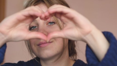 Portrait of a caucasian beautiful young woman 20 years old looking into the heart made gesture from her hands. A girl with blue eyes makes a heart gesture with her fingers. selective focus