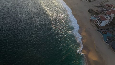 Drone shot of resorts on Playa El Médano with mountains in the distance in Cabo San Lucas Mexico, revealing and into the sun