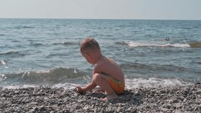 Cute baby boy playing with stones on the beach, on the seashore. Summer holidays and sea holidays. Authentic, slow-motion video