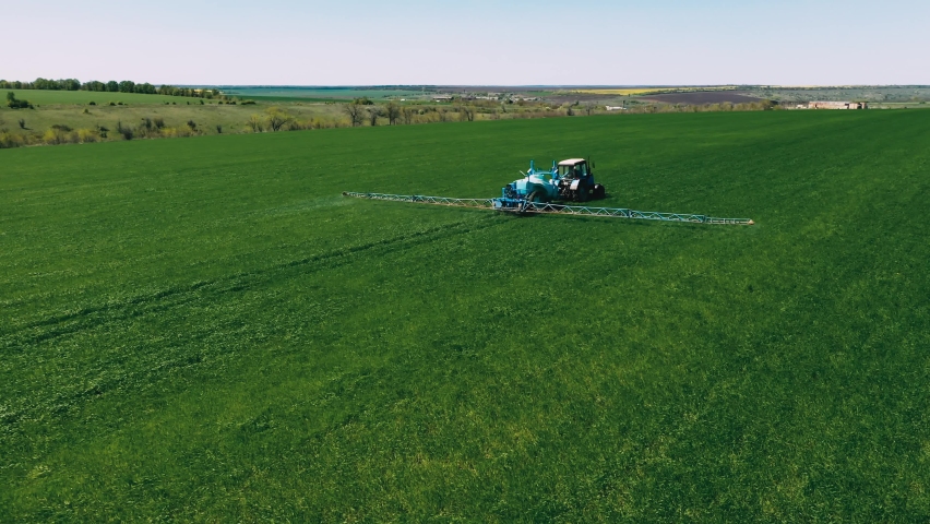 Spraying a green wheat field by tractor. The farming tractor spraying on field with sprayer, herbicides and pesticides. Industrial machine fertilizing a field. Chemicals used by agricultural tractor. | Shutterstock HD Video #1090092523