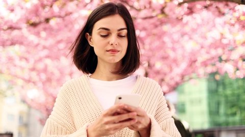 Close up attractive caucasian woman hold smartphone, watching social media standing on a city street near sakura trees. Smiling young woman texting on her phone with pink flower background.