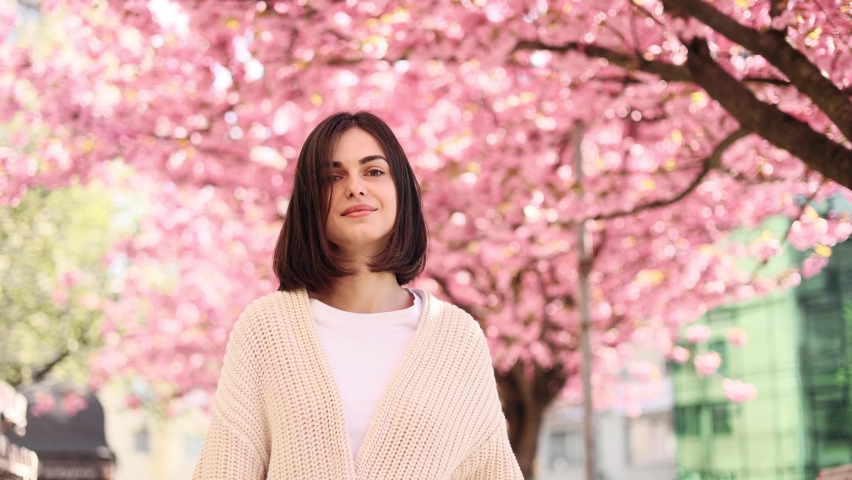 Charming young dark haired woman walking down the city street and enjoying beautiful day with sakura flowers in the background Pretty woman looking at the camera at spring morning outdoors Royalty-Free Stock Footage #1090093075