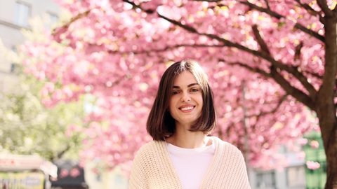 Portrait of a cute young woman while walking down the city street with sakura flowers in the background. Beautiful woman looking at the camera outside.