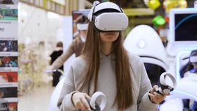 A beautiful young girl in a virtual reality headset draws abstract figures using two joystick controllers. A creative young girl is playing an augmented reality game against the background of other