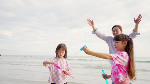 Happy Asian family on beach vacation. Mother with two little daughter walking and playing soap bubbles together on the beach at summer sunset. Mom and child girl kid enjoy outdoor activity lifestyle.