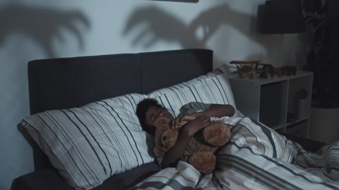 Medium of nine-year-old Black boy hugging toy bear, sleeping at home in dark, tossing and turning in his bed, having nightmare of monsters reaching for him